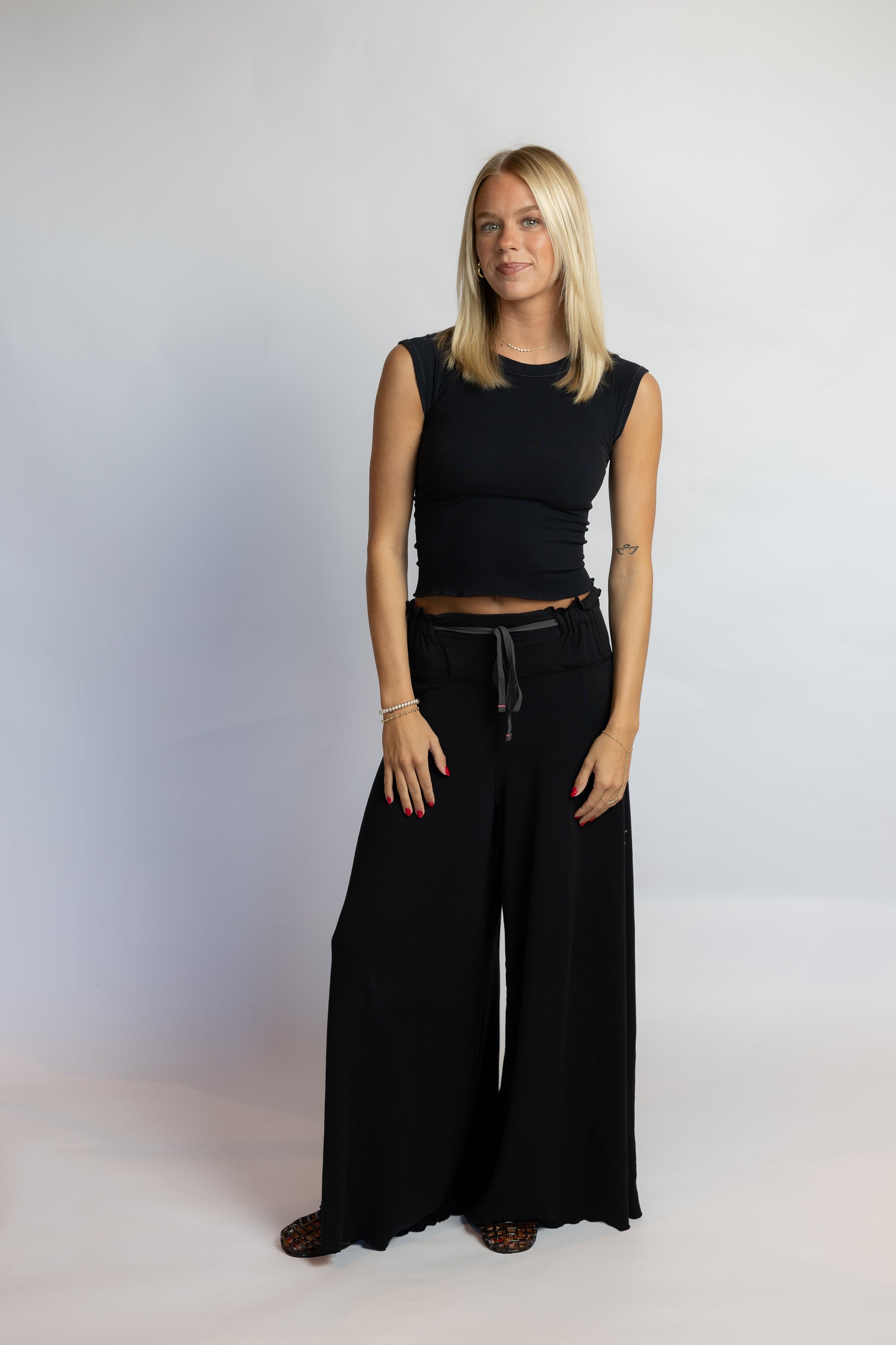A blonde woman modeling a black sleeveless crop top and matching wide-leg drawstring pants against a light gray studio backdrop.