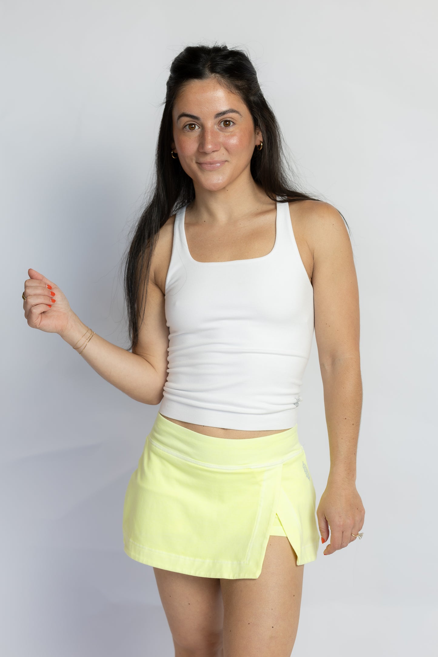 A woman modeling a white ribbed tank top and a neon yellow wrap-style athletic skirt against a light gray studio background.