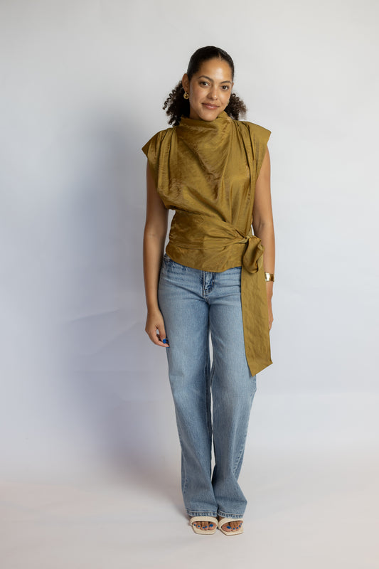 A woman modeling a sleeveless, draped olive green top with a high neck and side tie, paired with light-wash blue jeans and beige sandals, standing against a light gray studio backdrop.