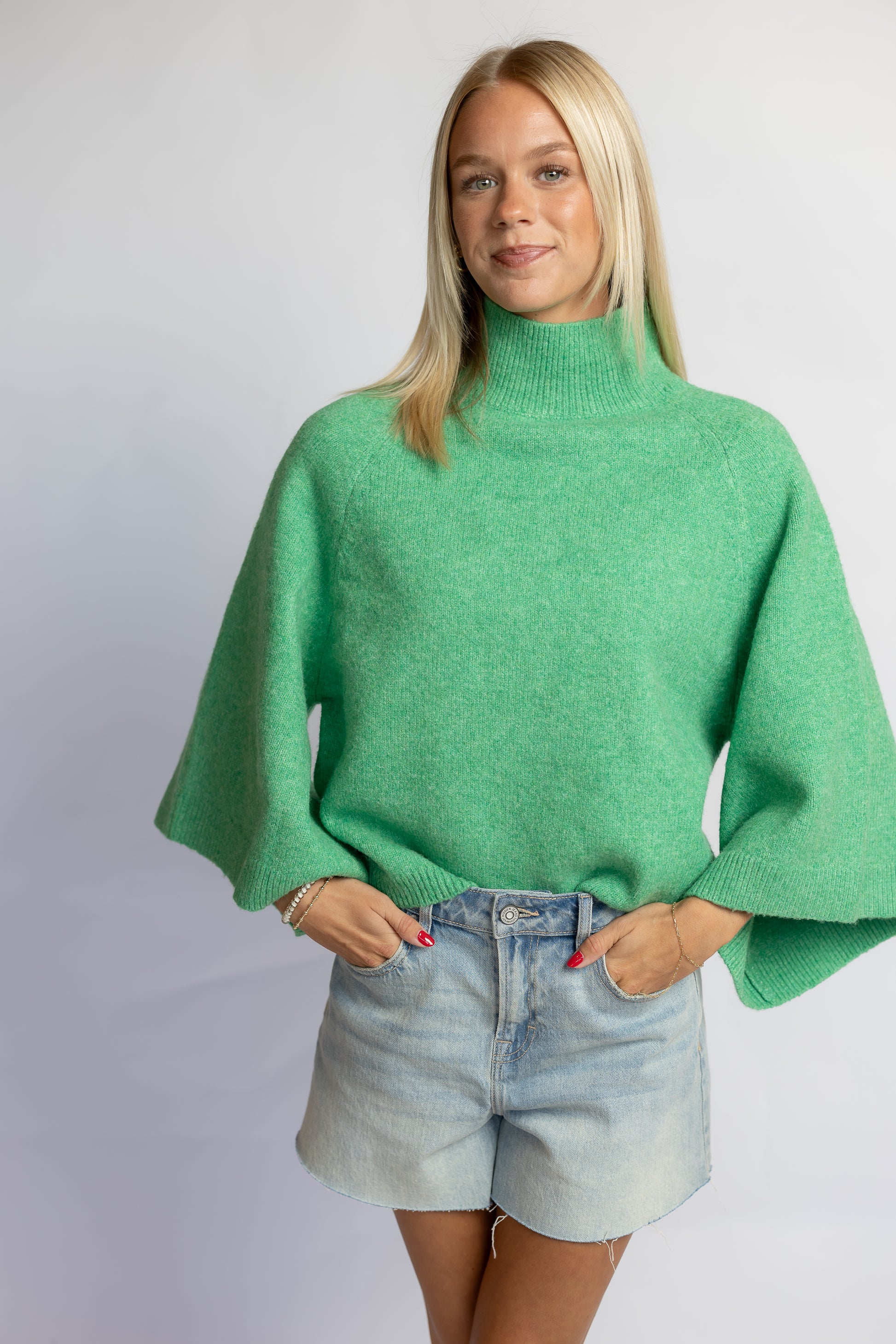 A woman modeling a bright green knit turtleneck sweater with wide, draped sleeves, paired with light-wash denim shorts, against a plain light gray studio background.