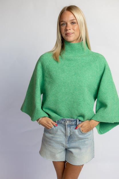 A woman modeling a bright green knit turtleneck sweater with wide, draped sleeves, paired with light-wash denim shorts, against a plain light gray studio background.