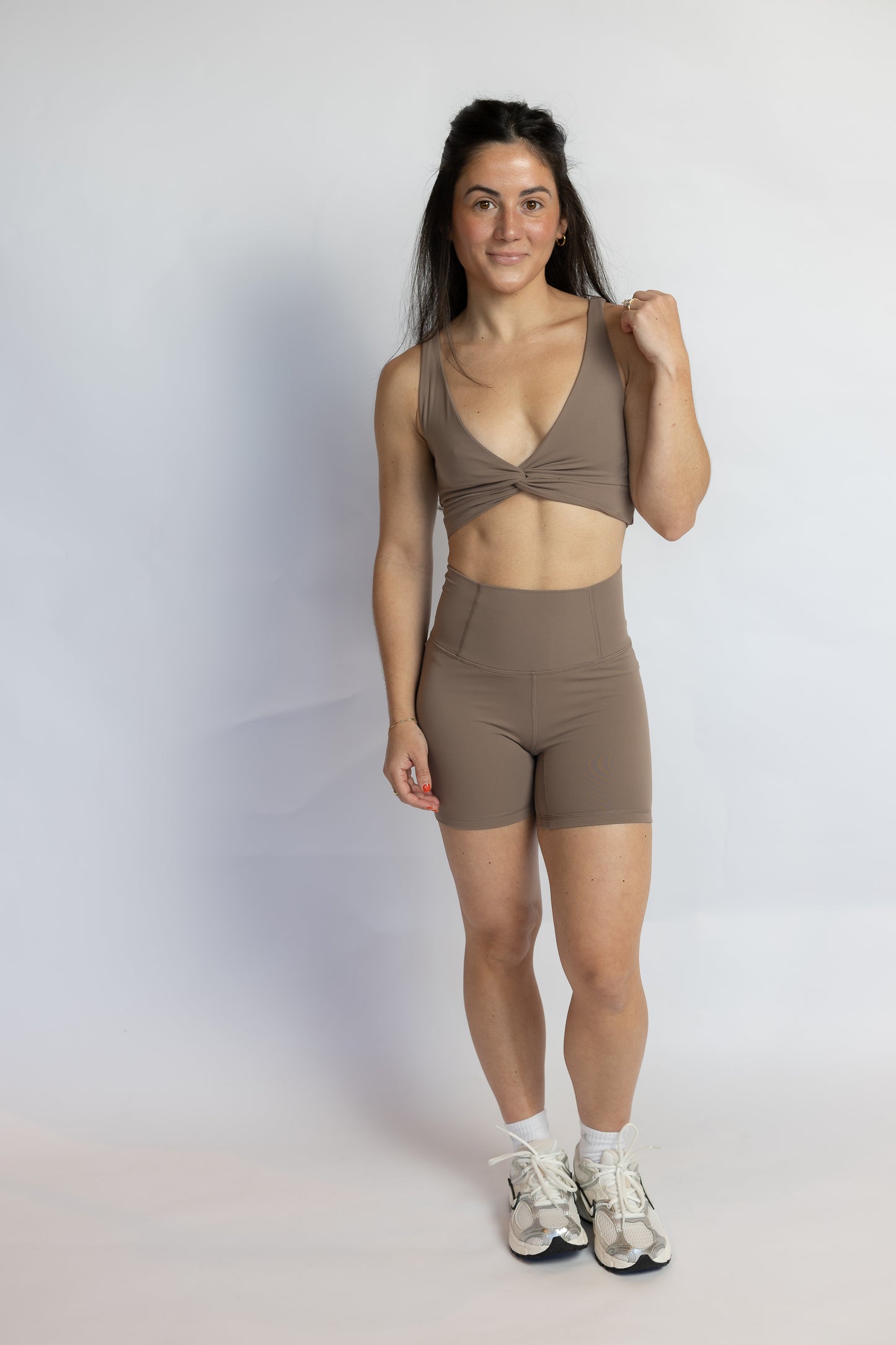A woman modeling a matching taupe-brown activewear set consisting of a crossover-front sports bra and high-waisted shorts, paired with white socks and athletic sneakers, against a light gray studio background.
