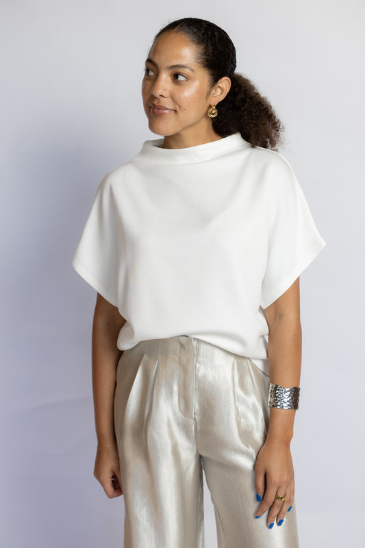 A woman wearing a white short-sleeve mock-neck top and shiny light beige trousers, standing against a light gray studio background.