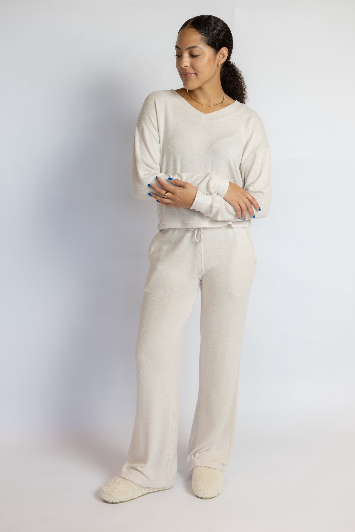 A woman modeling a matching cream-colored loungewear set consisting of a long-sleeve V-neck top and wide-leg drawstring pants, paired with matching slippers, against a plain light gray studio background.