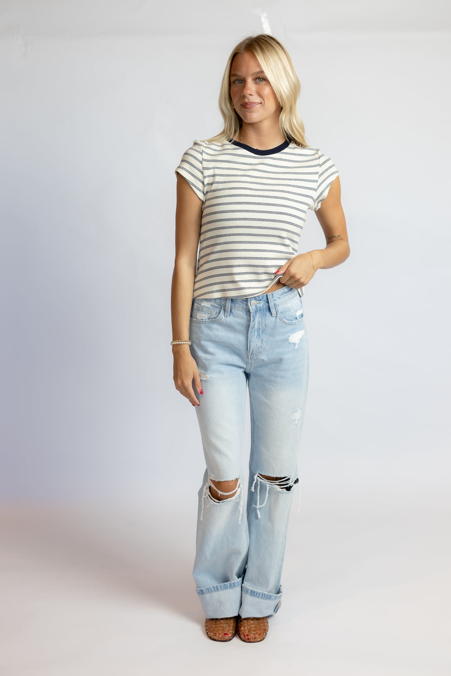 A blonde woman modeling a short-sleeve striped t-shirt with dark navy trim and light-wash distressed jeans, standing against a plain light gray studio background.