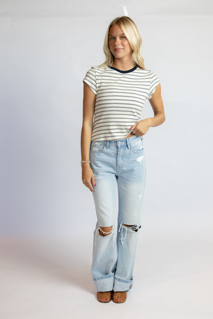 A blonde woman modeling a short-sleeve striped t-shirt with dark navy trim and light-wash distressed jeans, standing against a plain light gray studio background.