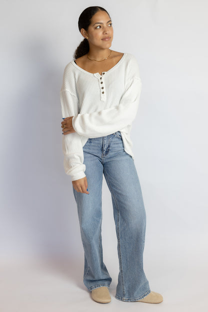 A woman modeling a white ribbed long-sleeve henley top with button details and light-wash blue wide-leg jeans, standing against a plain light gray studio background.
