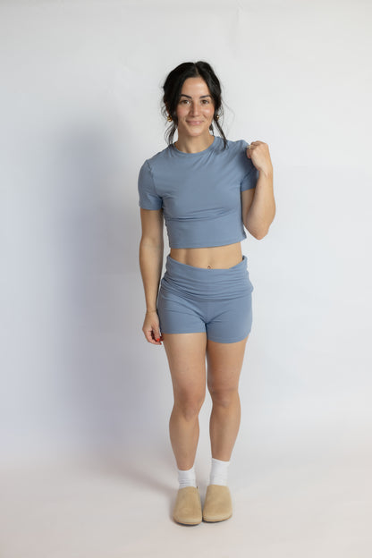 A woman modeling a matching dusty blue two-piece activewear set consisting of a short-sleeve crop top and high-waisted shorts, paired with white socks and beige slippers against a plain white studio background.