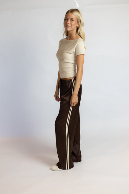 A blonde woman stands in profile wearing a fitted cream short-sleeve top and dark brown wide-leg pants with two vertical cream stripes down the sides, against a plain light gray studio background.