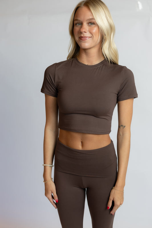 A blonde woman modeling a matching brown short-sleeve crop top and high-waisted leggings set against a light gray studio background.
