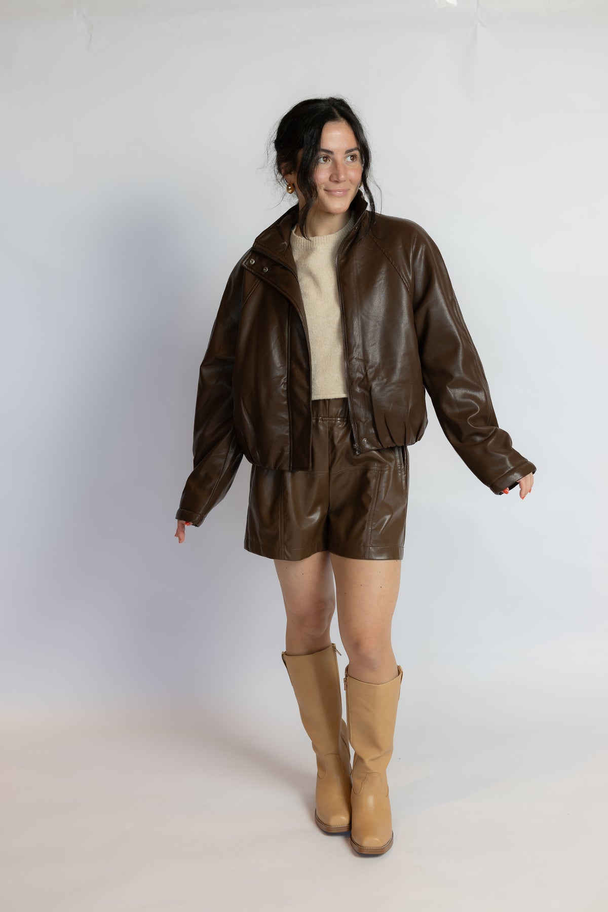 A model stands against a light gray studio background wearing a dark brown faux-leather jacket and matching shorts over a cream top, paired with tan knee-high boots.