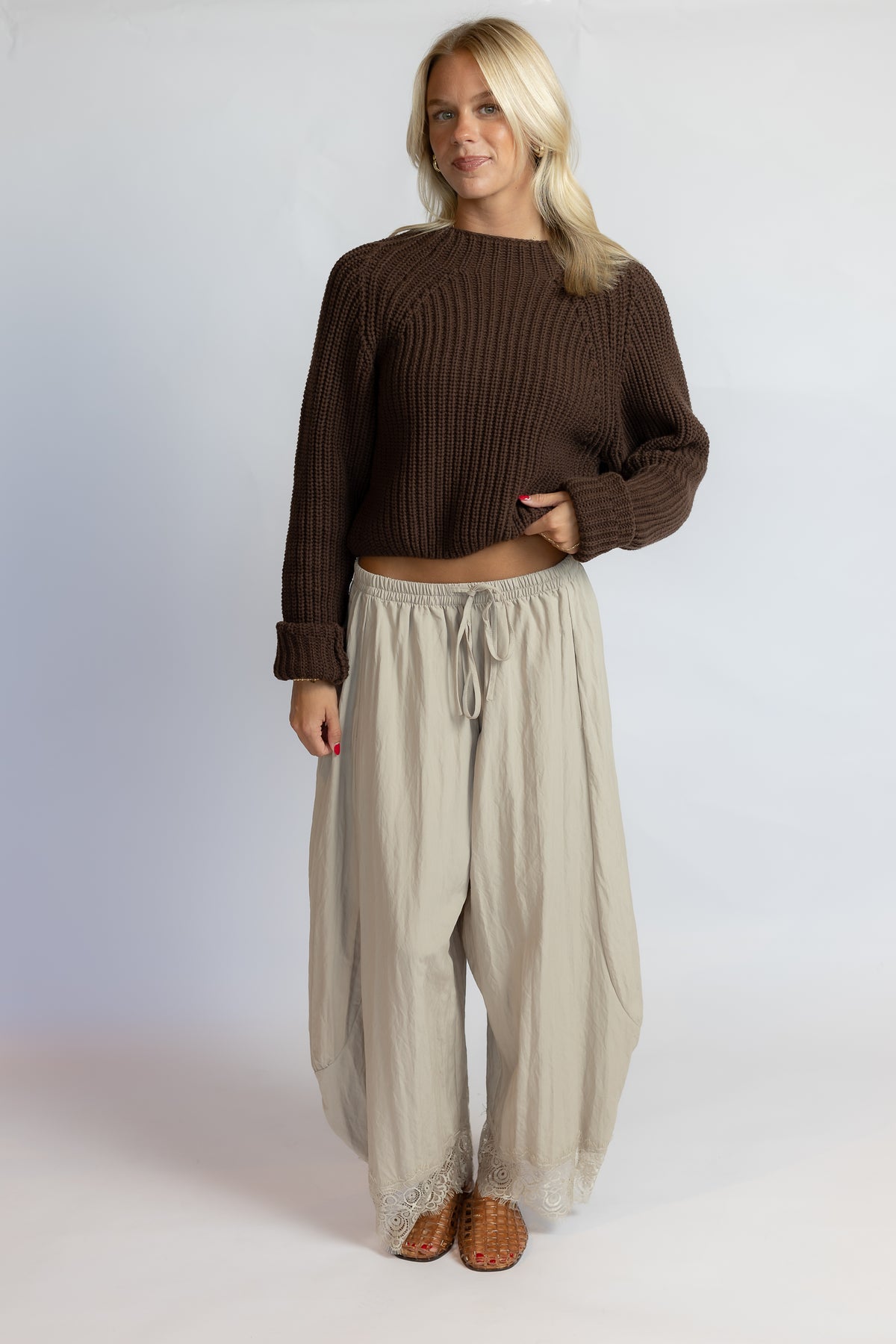 A woman modeling a dark brown ribbed knit cropped sweater and light beige wide-leg drawstring pants against a light gray studio background.