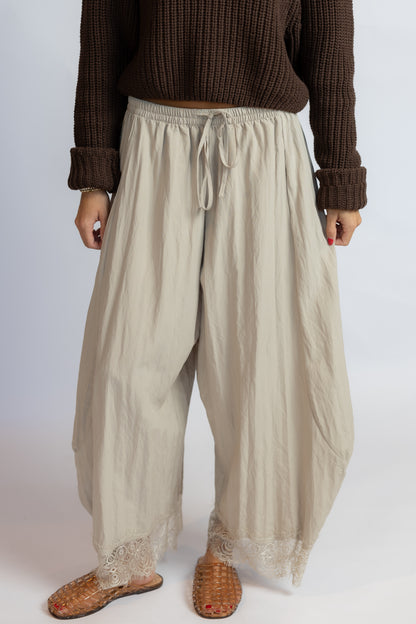 A person wearing loose, wide-leg beige drawstring pants with lace trim at the hem, paired with a brown ribbed sweater and woven brown shoes, shown against a plain light background.