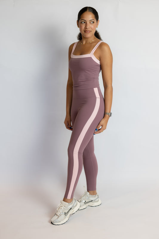 A woman modeling a matching mauve athletic set consisting of a tank top and high-waisted leggings with light pink side panels, paired with white Nike running shoes, against a light gray studio background.
