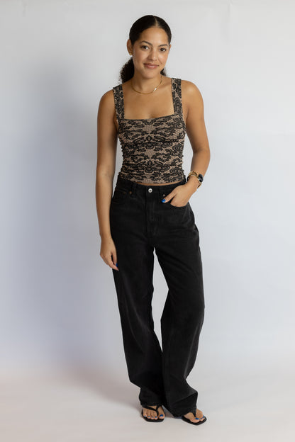 A woman modeling a black lace sleeveless top with a square neckline and black wide-leg jeans, standing against a light gray studio background.