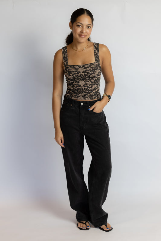 A woman modeling a black lace sleeveless top with a square neckline and black wide-leg jeans, standing against a light gray studio background.
