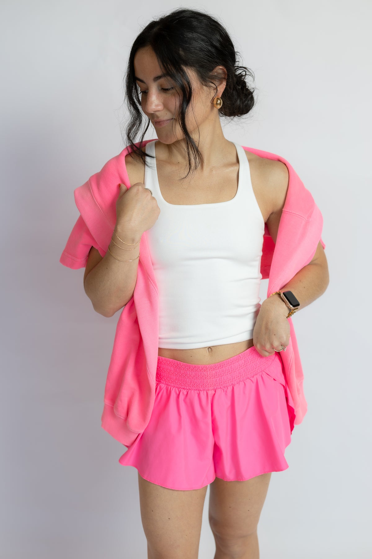A woman modeling a bright pink athletic set consisting of a tank top, matching shorts, and an open zip-up hoodie, paired with a white tank top underneath, against a plain light gray background.