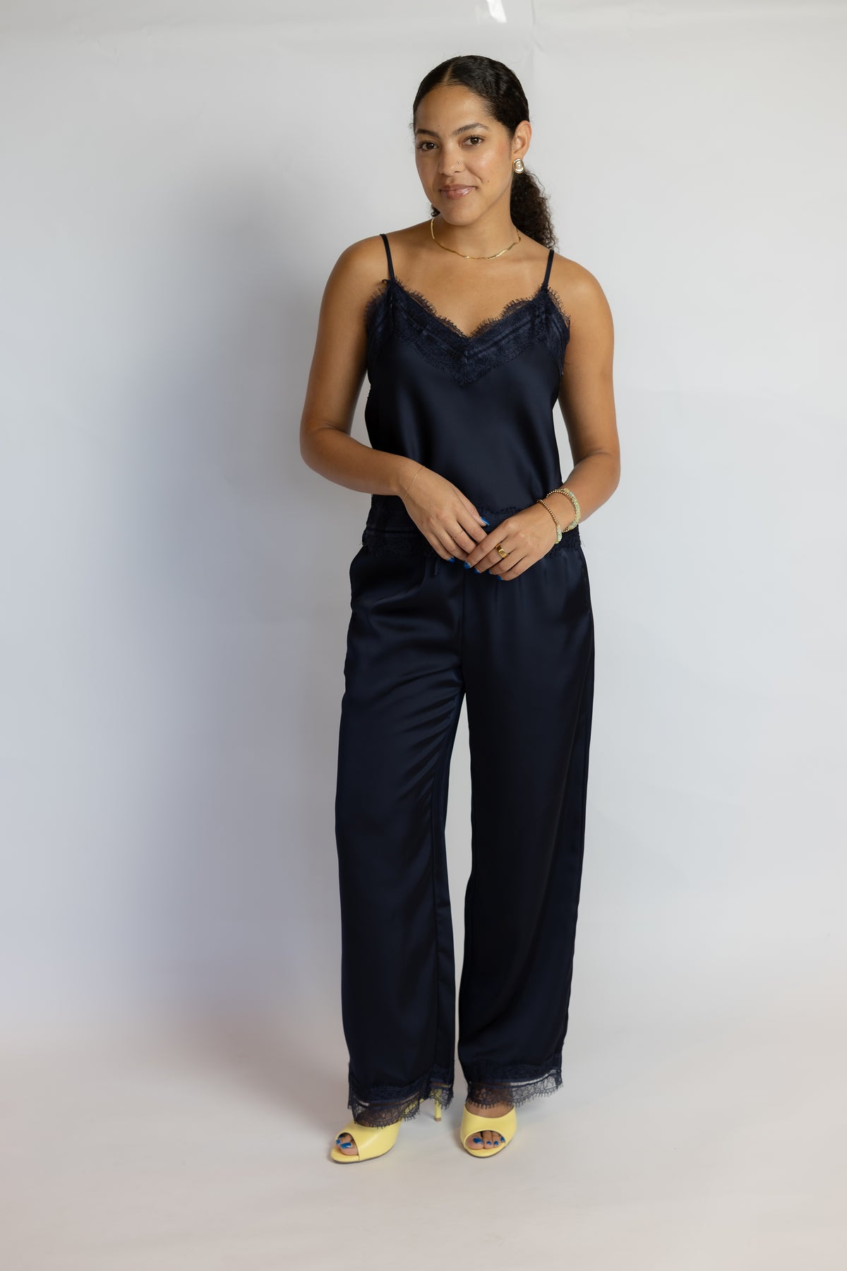 A woman modeling a dark navy blue satin pajama set consisting of a camisole top with lace trim and matching wide-leg pants, paired with yellow slide sandals, against a plain white studio background.