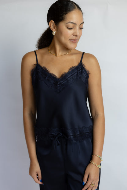 A woman wearing a dark navy blue satin camisole top with lace trim and matching satin pants, photographed against a plain light gray studio background.