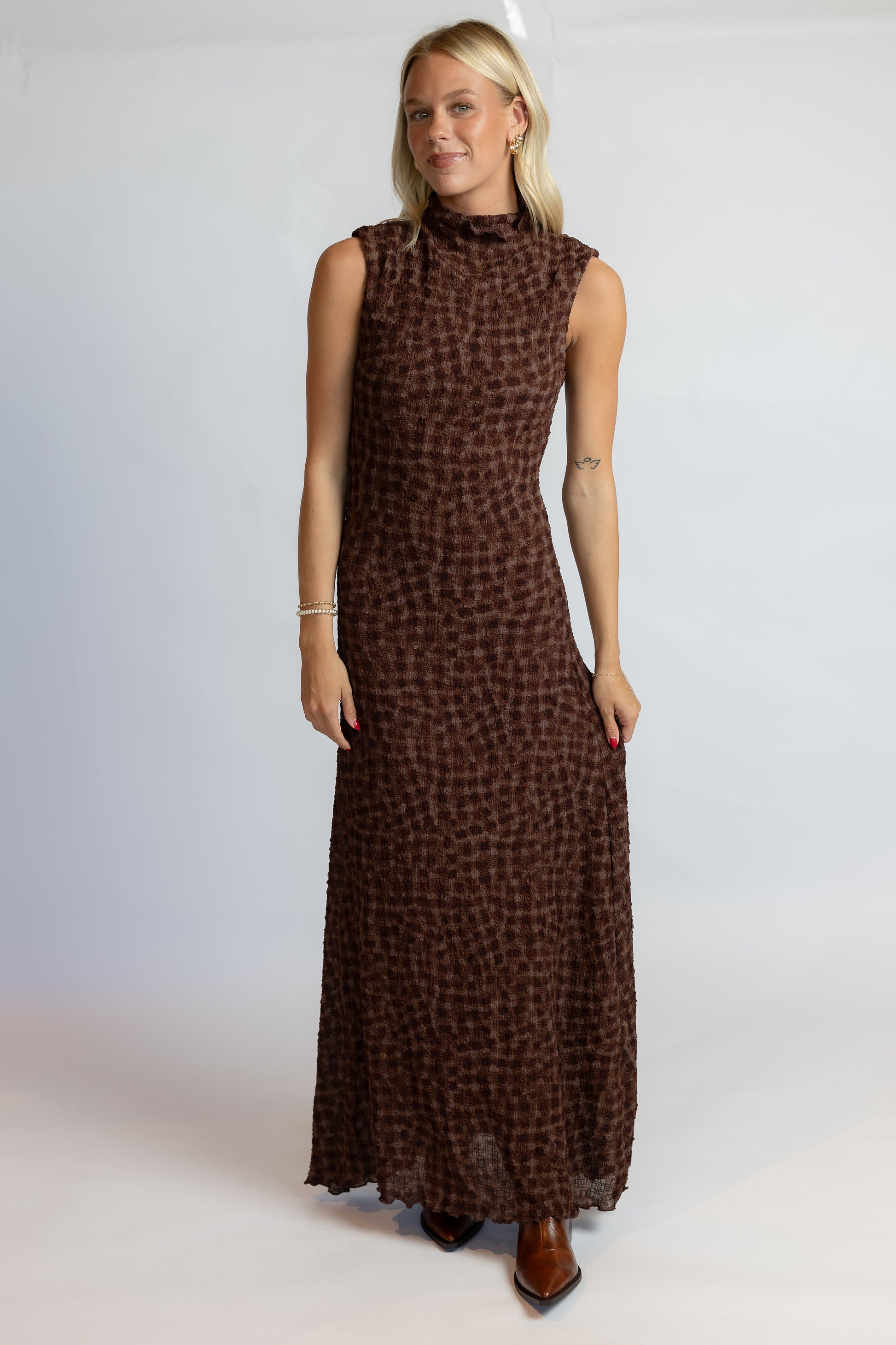 A woman modeling a sleeveless, high-neck, floor-length brown dress with a small-scale leopard-like print, set against a plain light gray studio background.