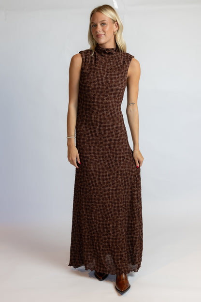 A woman modeling a sleeveless, high-neck, floor-length brown dress with a small-scale leopard-like print, set against a plain light gray studio background.