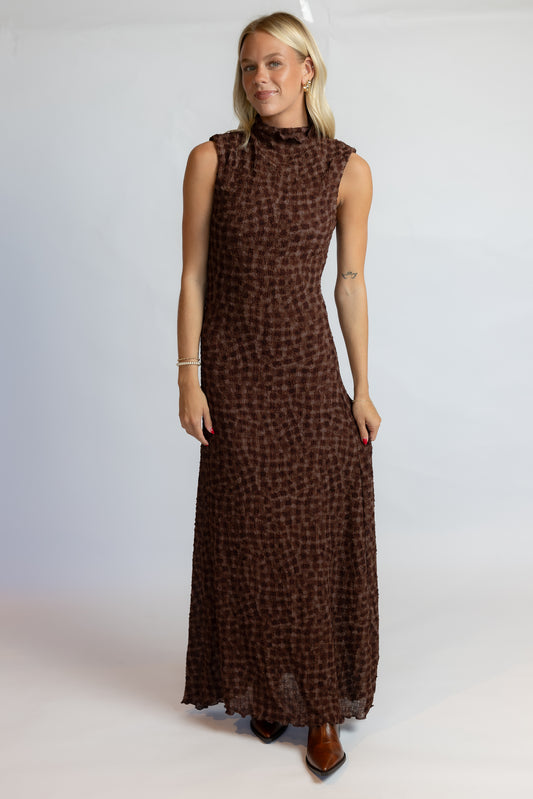 A woman modeling a sleeveless, high-neck, floor-length brown dress with a small-scale leopard-like print, set against a plain light gray studio background.