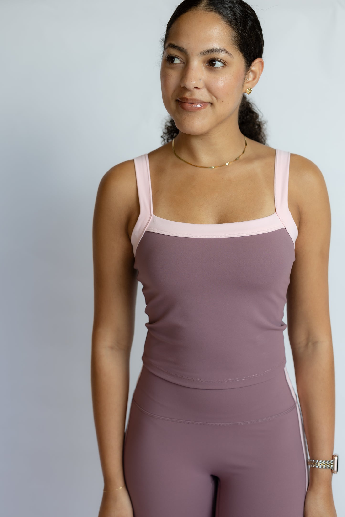 A woman modeling a mauve-colored activewear set consisting of a square-neck tank top and matching leggings, featuring light pink trim on the straps and neckline, set against a plain light gray background.