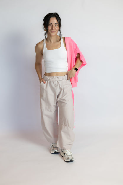 A woman modeling a white ribbed tank top, beige wide-leg cargo pants, and white sneakers, with a bright pink jacket draped over her shoulder, against a plain white studio background.