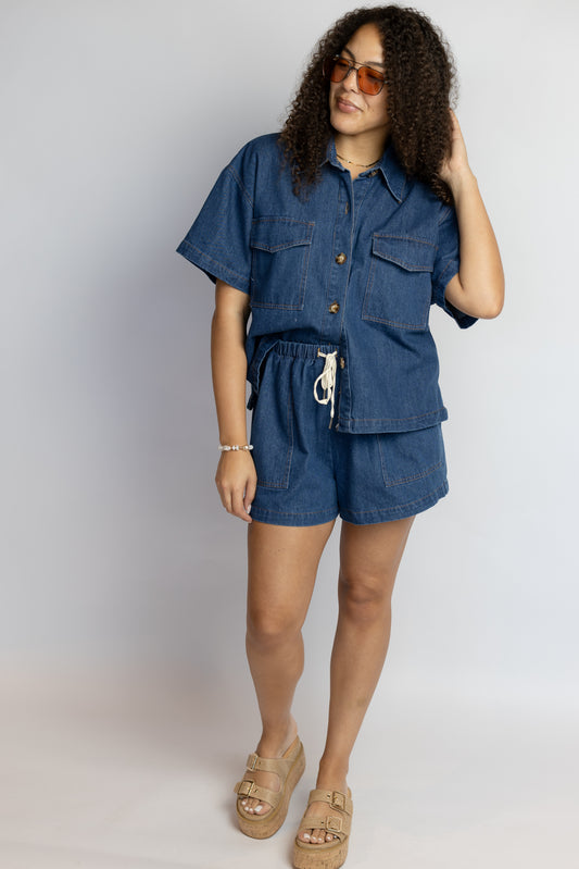 Woman wearing a blue denim set outfit with sunglasses on a plain background