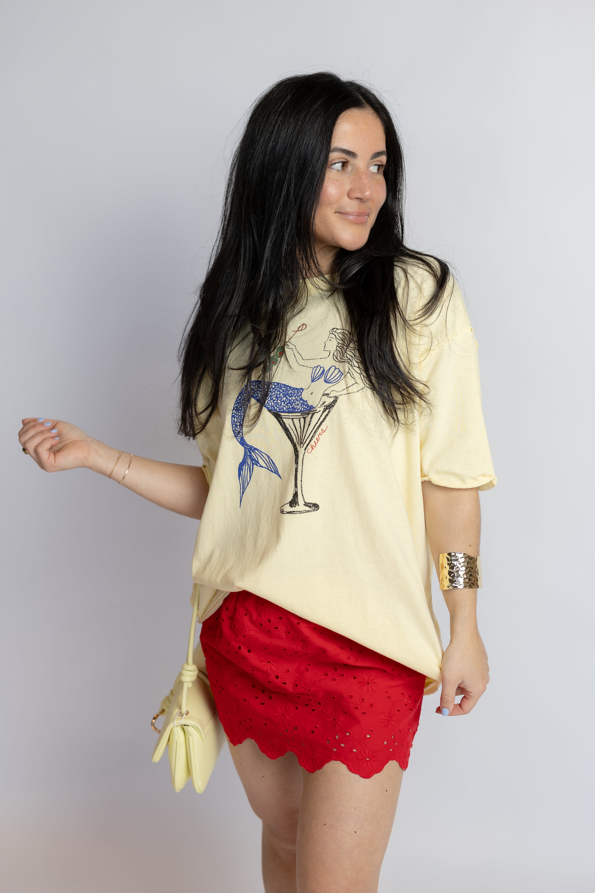 Woman wearing a yellow t-shirt with a graphic design and red shorts, holding a yellow bag against a plain background.