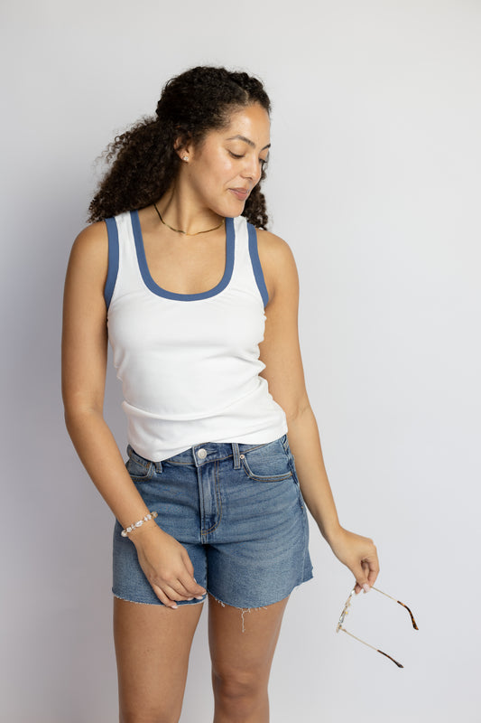 Woman wearing a white ribbed scoop neck tank top with blue contrast trim and denim shorts.