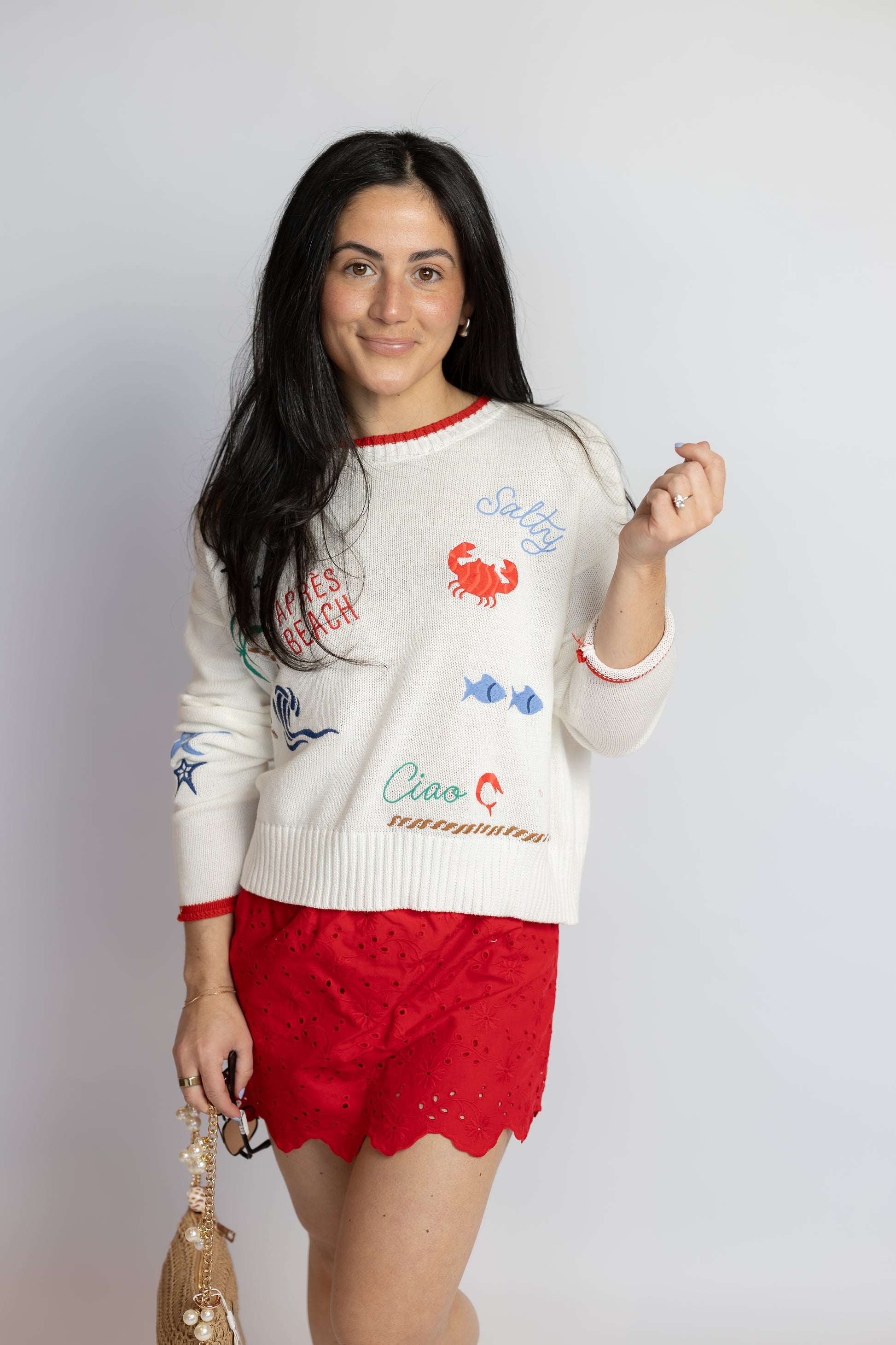 Woman wearing a lightweight cream cotton sweater with colorful coastal-inspired embroidery and red trim details.