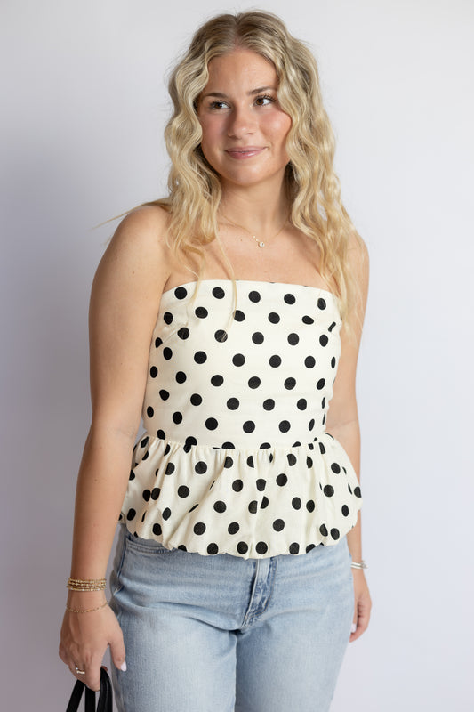 Woman wearing a cream strapless bubble hem top with black polka dots paired with light wash denim jeans.