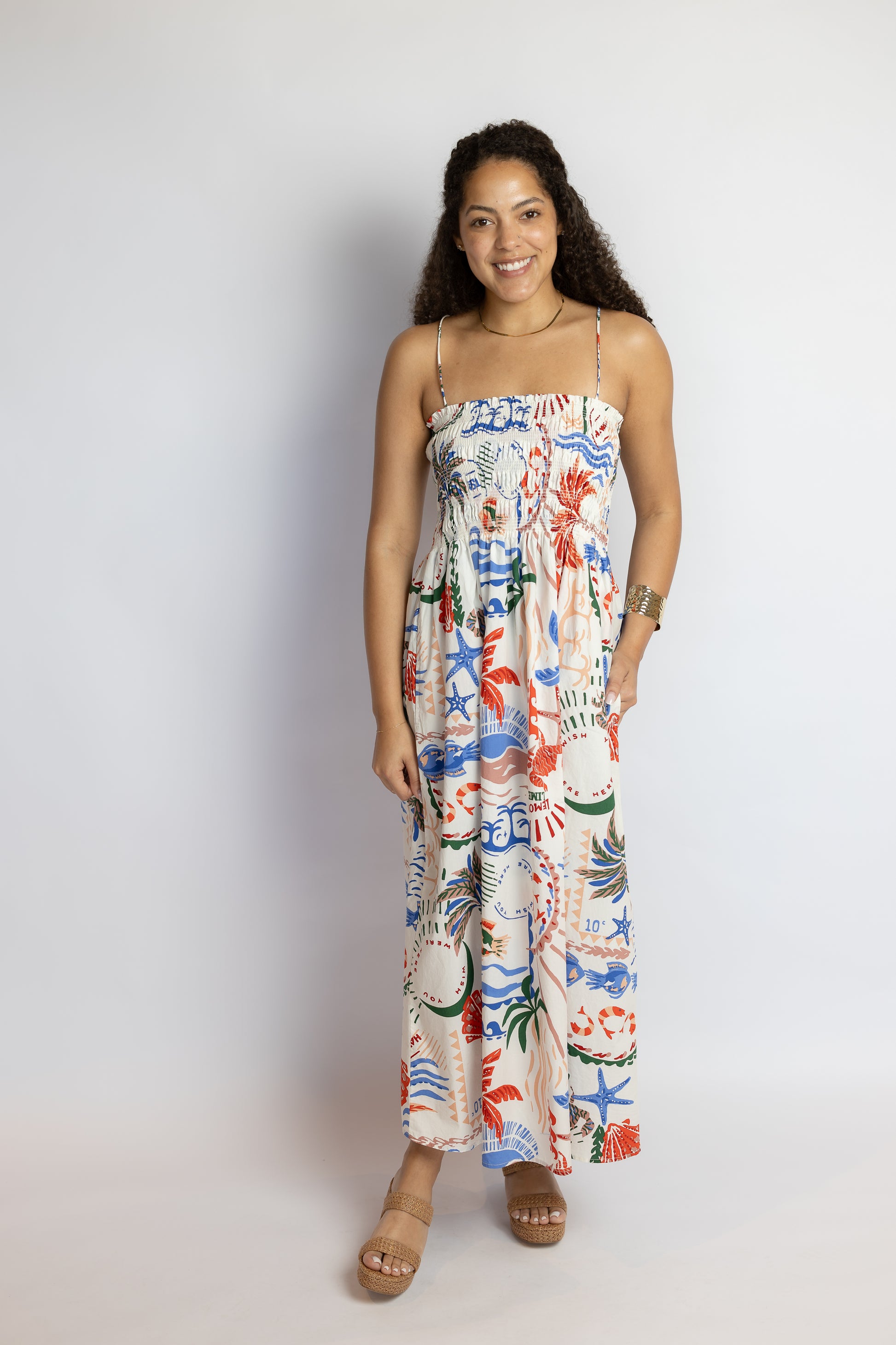 Woman wearing a coastal print midi dress with a smocked bodice, spaghetti straps, and colorful beach-inspired motifs.