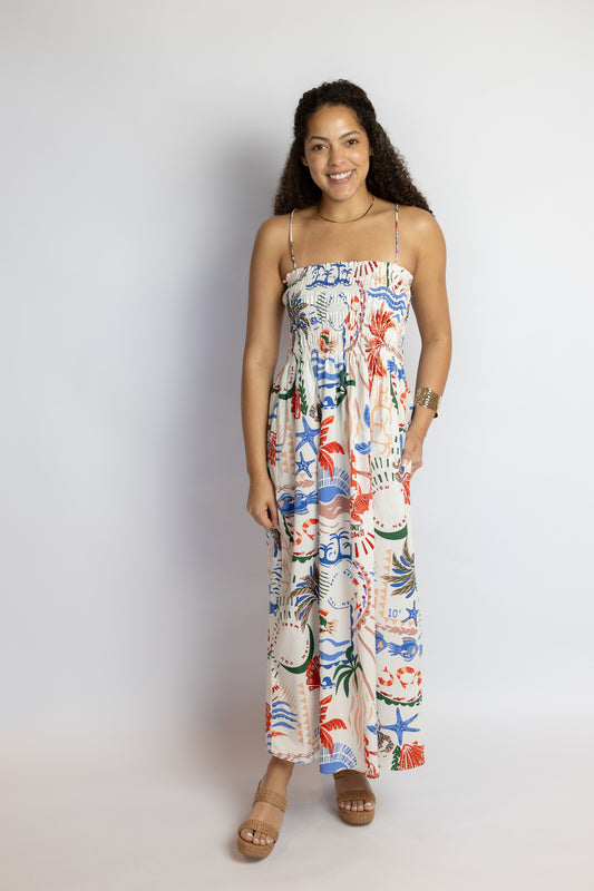 Woman wearing a coastal print midi dress with a smocked bodice, spaghetti straps, and colorful beach-inspired motifs.