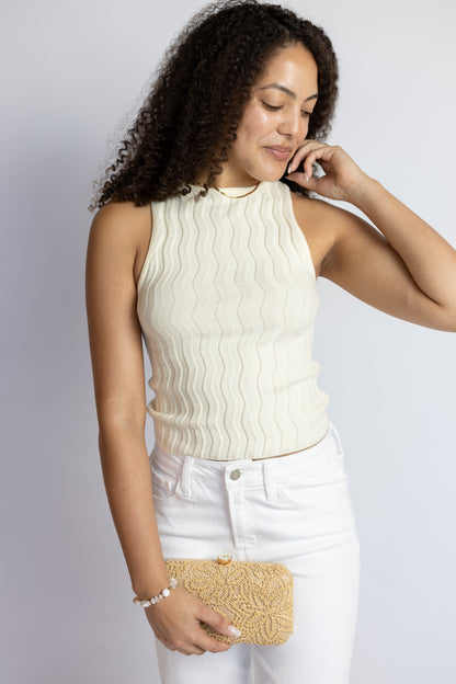Woman wearing a cream sleeveless textured sweater top with a boat neckline and white denim jeans.