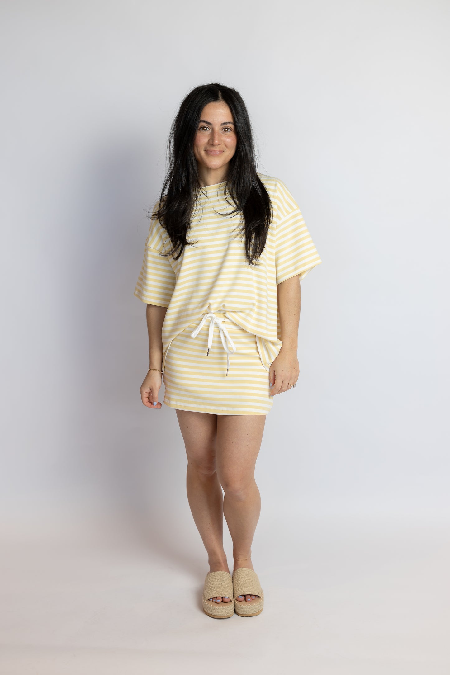 Woman wearing an oversized yellow and white striped t-shirt styled with a matching striped mini skirt and woven slide sandals.