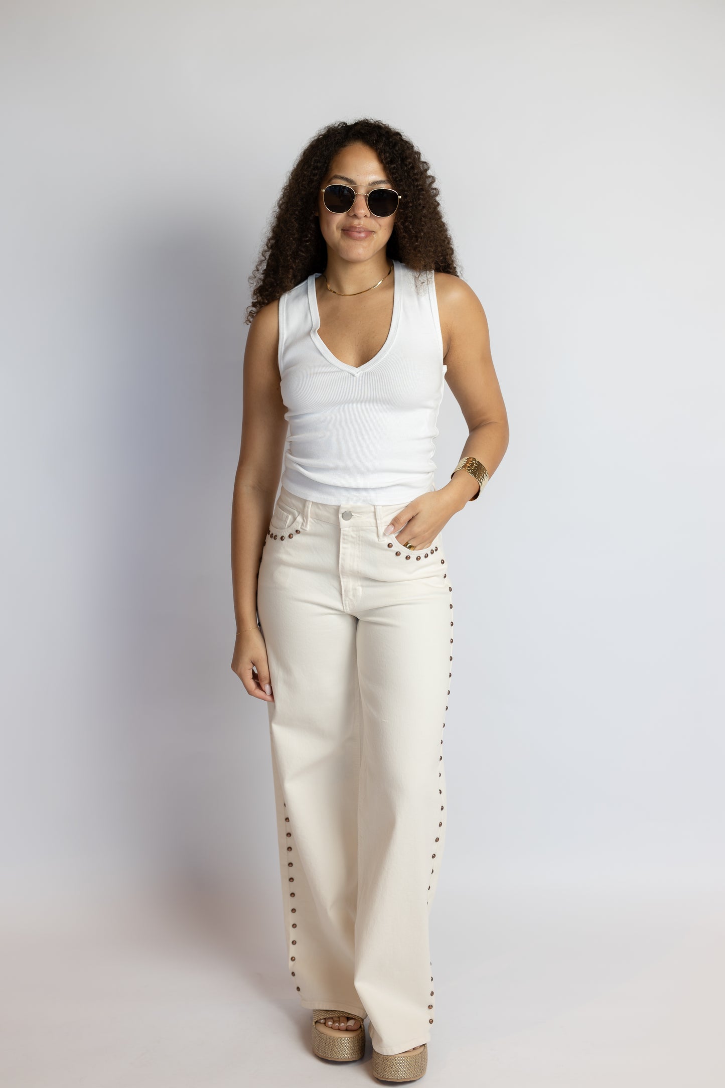 Woman wearing a white sleeveless top and beige pants with studded details against a plain background