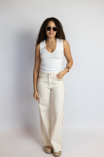 Woman wearing a white sleeveless top and beige pants with studded details against a plain background