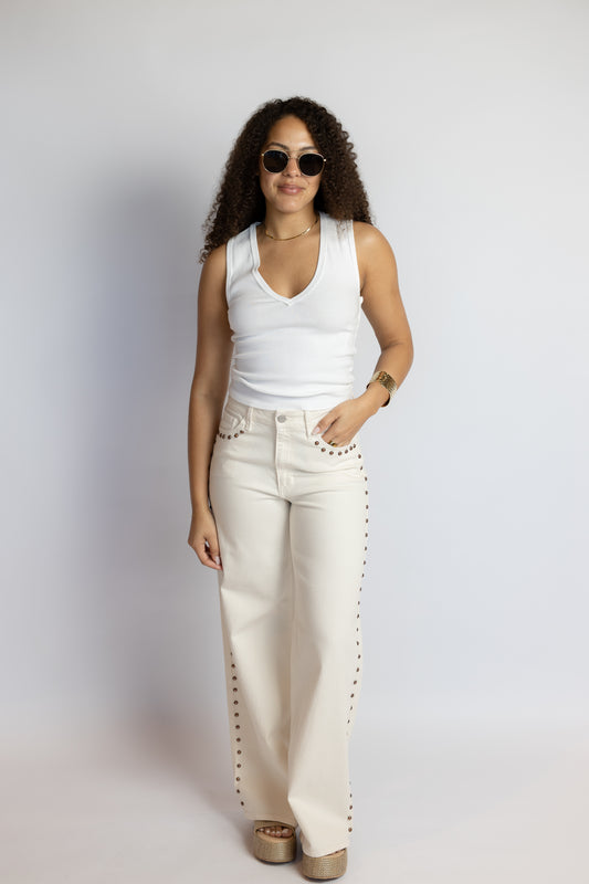 Woman wearing a white sleeveless top and beige pants with studded details against a plain background