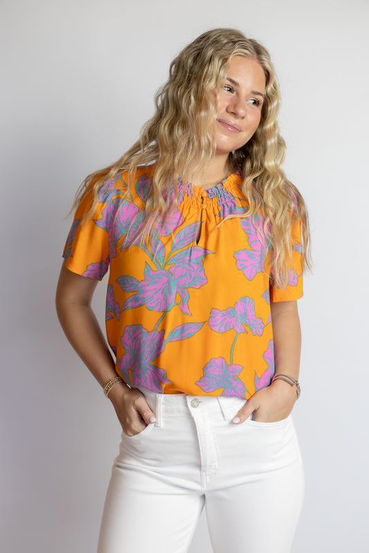 Woman wearing a vibrant orange floral blouse with pink flowers, smocked neckline, and white denim jeans.