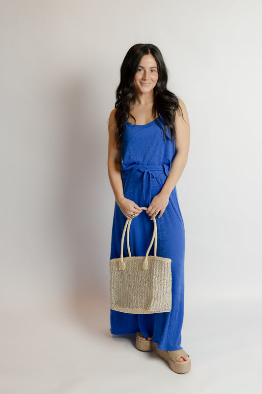 Woman in a blue dress holding a straw bag against a plain background