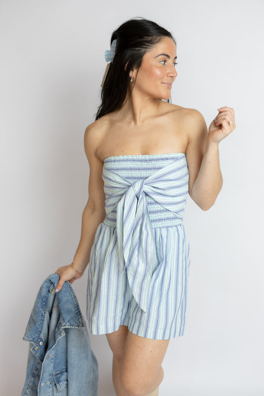 Woman wearing a strapless blue and white striped romper holding a denim jacket against a plain background