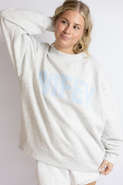 Woman wearing a light gray sweatshirt with 'WIFEY' printed on it against a white background