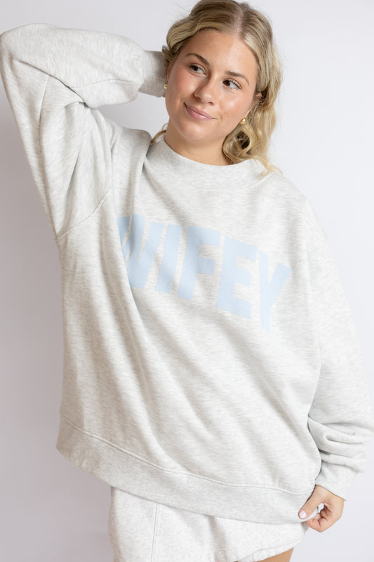 Woman wearing a light gray sweatshirt with 'WIFEY' printed on it against a white background