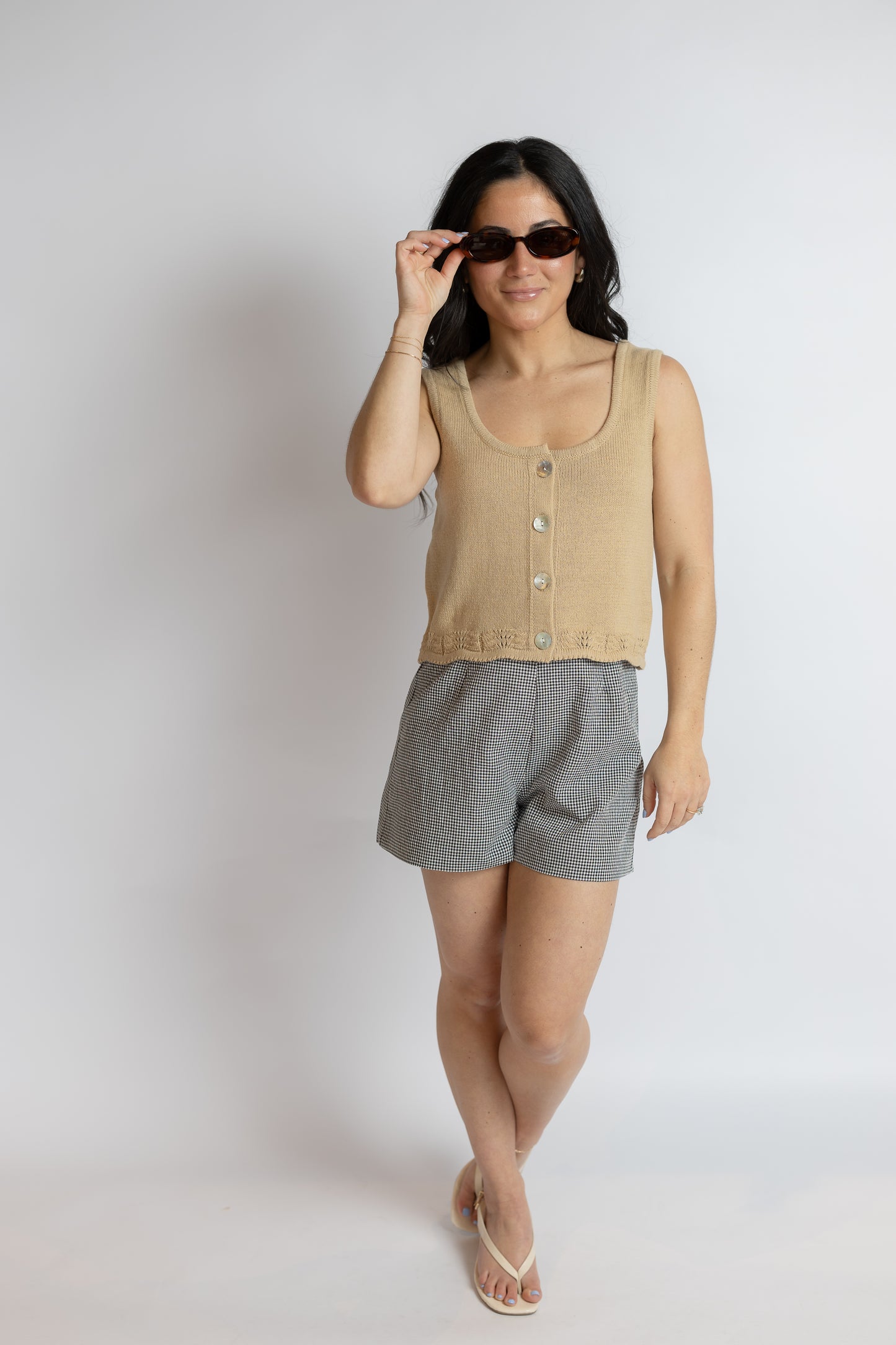 Woman wearing a beige sleeveless top and gray shorts, holding sunglasses against a plain background