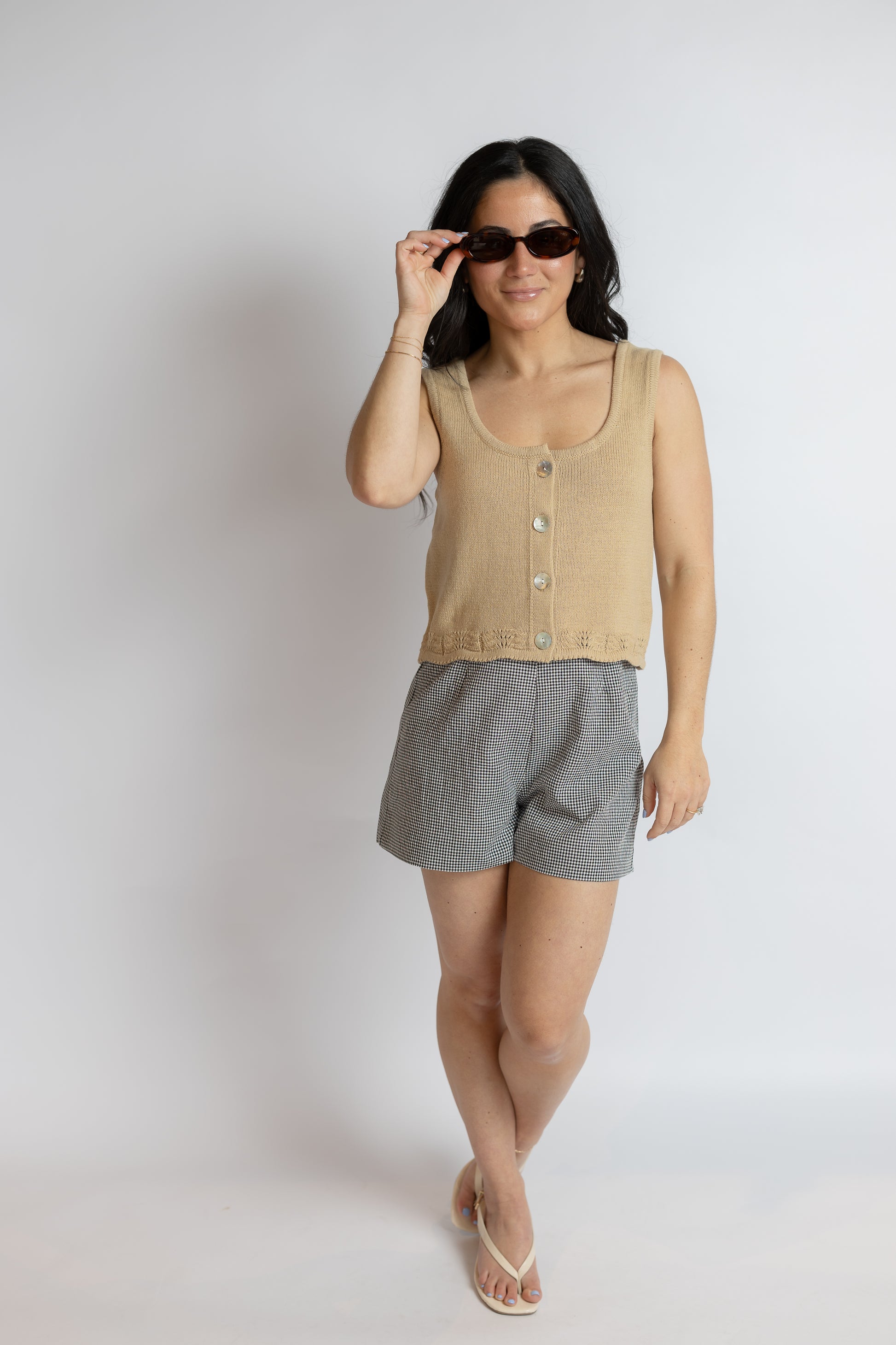 Woman wearing a beige sleeveless top and gray shorts, holding sunglasses against a plain background