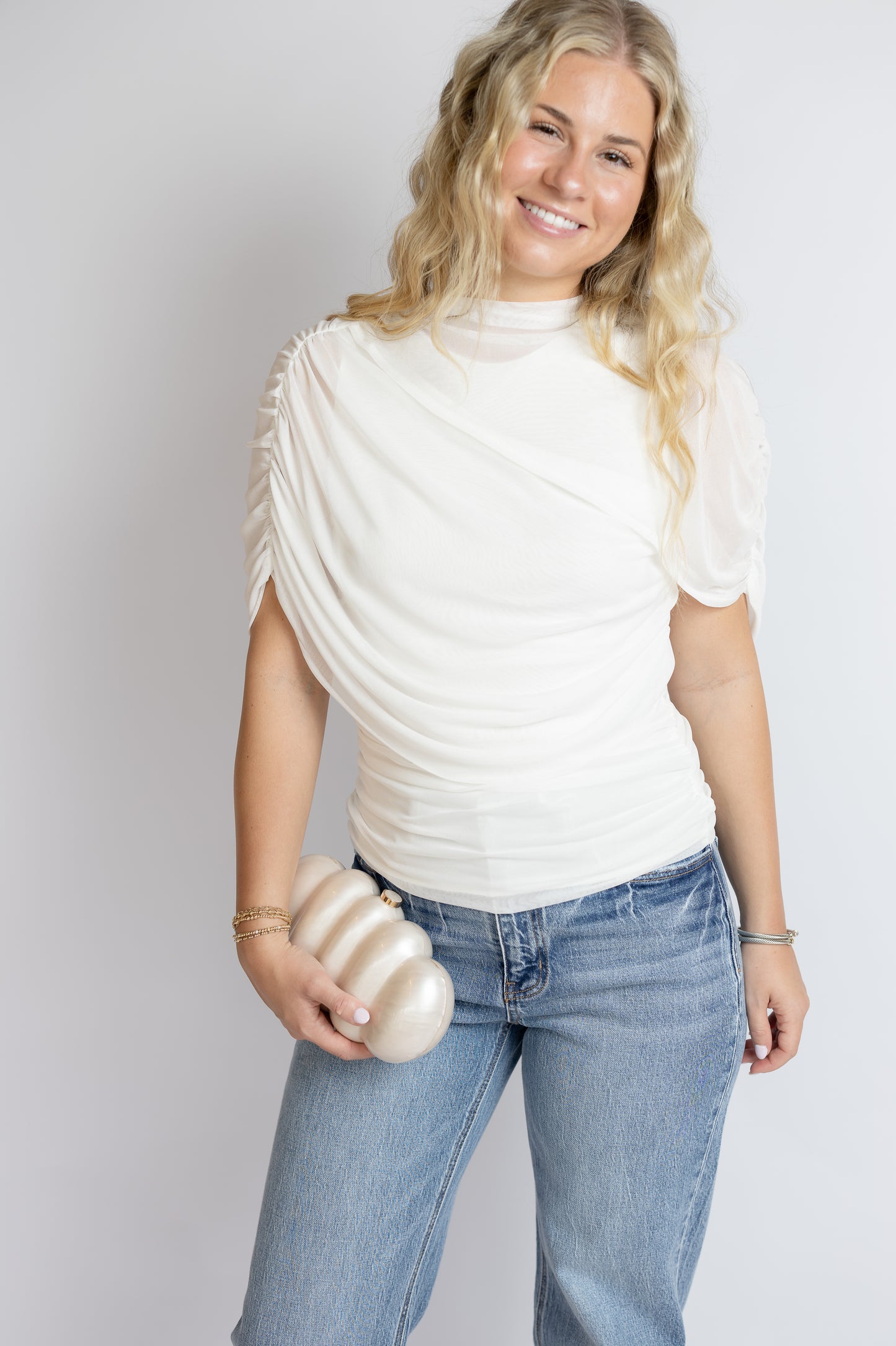 Woman wearing a white bridal ruched top and blue jeans holding a marble purse against a plain background