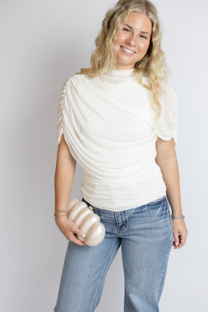 Woman wearing a white bridal ruched top and blue jeans holding a marble purse against a plain background