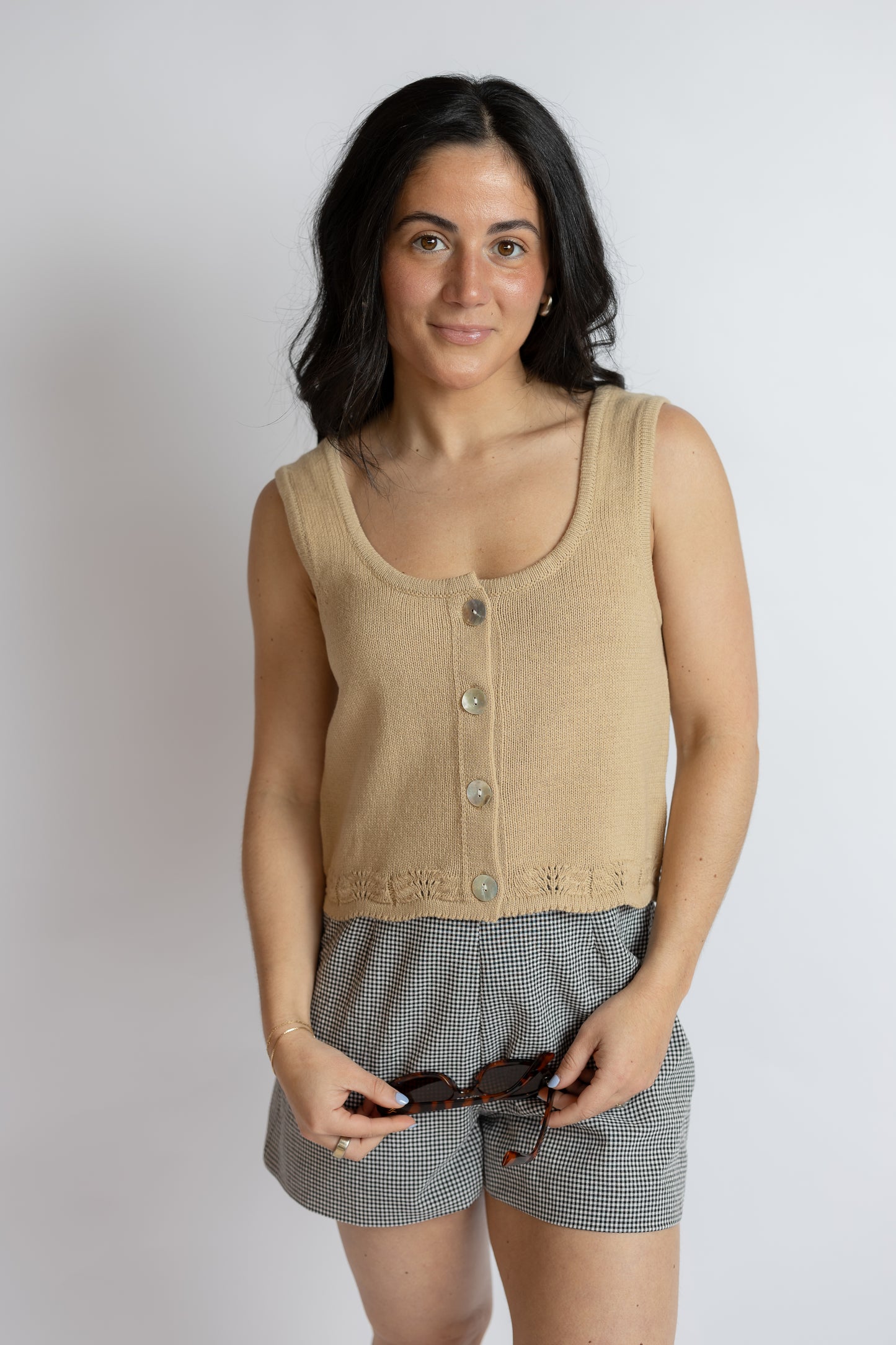 Woman wearing a beige sleeveless sweater button down top and gray checkered shorts against a plain background